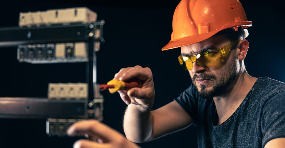 A male electrician in a protective helmet works in a switchboard with an electrical connecting cable.